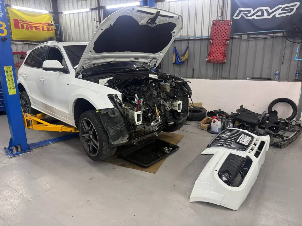 Audi Q5 2014: На фото белый Audi Q5 2014 года, поднятый на подъёмнике. Передняя часть кузова снята, виден разобранный двигательный отсек, снятый бампер лежит на полу. Работа ведётся в автосервисе.