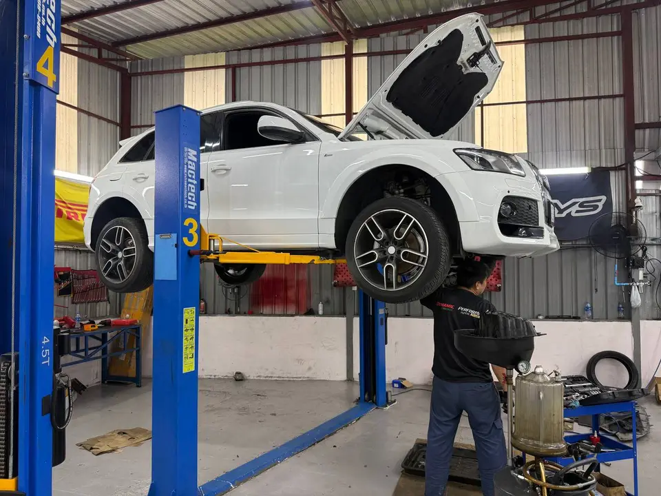 Audi Q5 2014: Белый Audi Q5 поднят на подъёмнике, капот открыт. Работник в гараже стоит у автомобиля, рядом видны инструменты и запчасти.