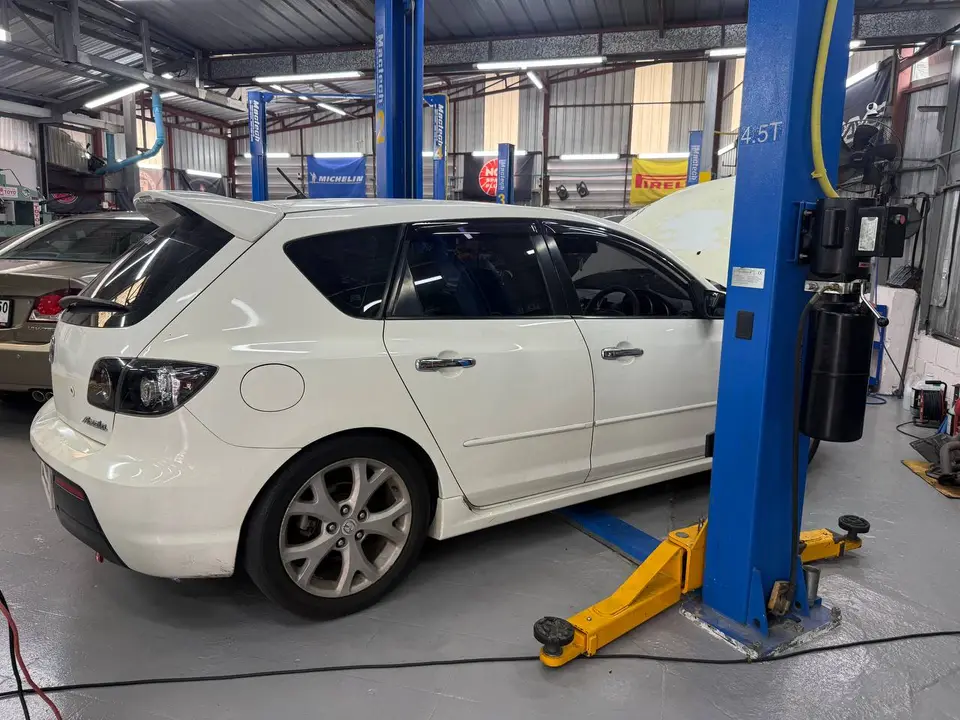 Mazda 3 2013: Белый Mazda 3 2013 года поднят на подъёмнике в автосервисе. На фото видна задняя часть автомобиля, подъёмник, инструменты и оборудование в гараже.
