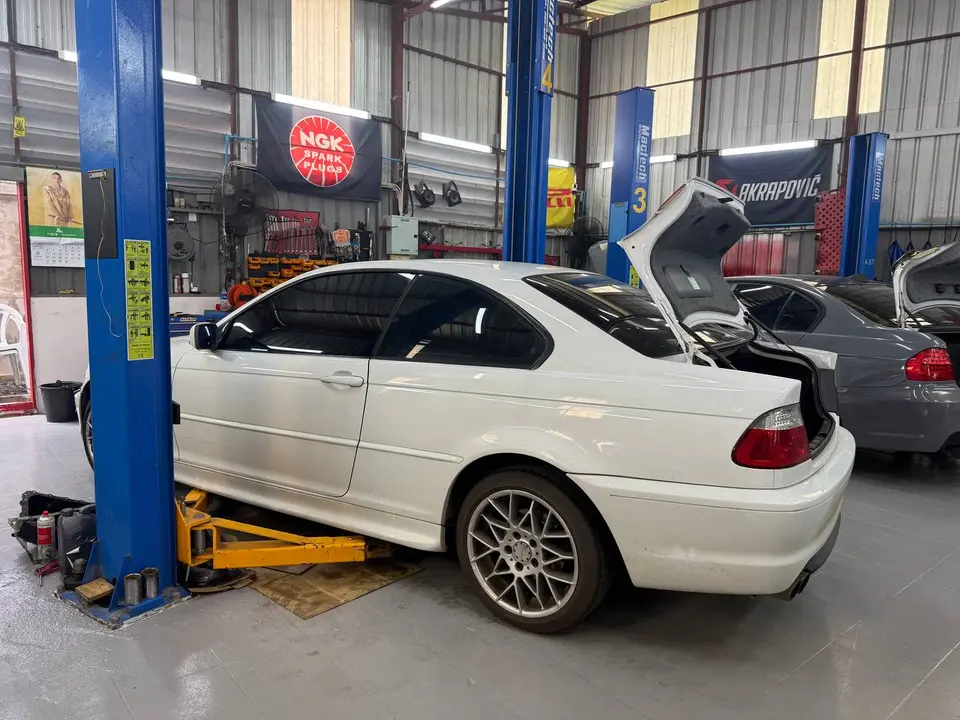 BMW 3 2010: Белый BMW 3-й серии 2010 года поднят на подъёмнике в автосервисе. Открыты багажник и капот, видны инструменты и оборудование для обслуживания.