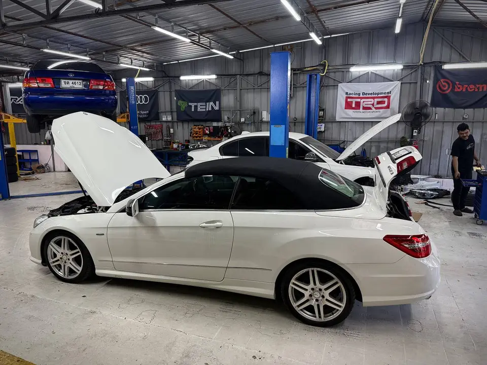 Mercedes-Benz E250 2015: На фото белый Mercedes-Benz E250 кабриолет с открытым капотом и снятой мягкой крышей. Автомобиль находится в гараже, рядом с ним — другие машины на подъёмниках и техническое о