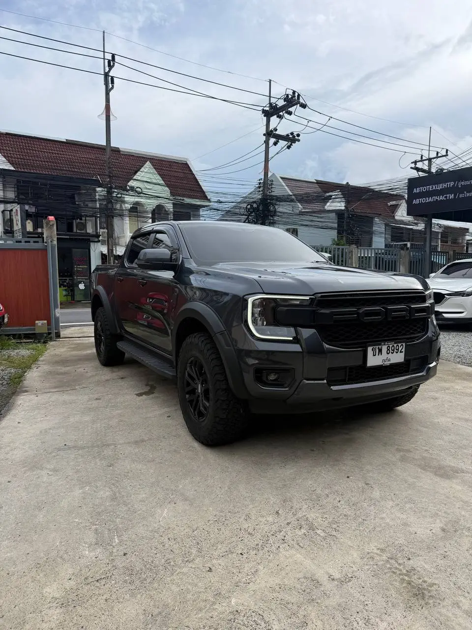 Ford Ranger 2025: На фото чёрный Ford Ranger 2025, стоящий на бетонной площадке. Видна передняя часть автомобиля с решёткой радиатора, фарами и номерным знаком. На заднем плане — жилые дома и провода.