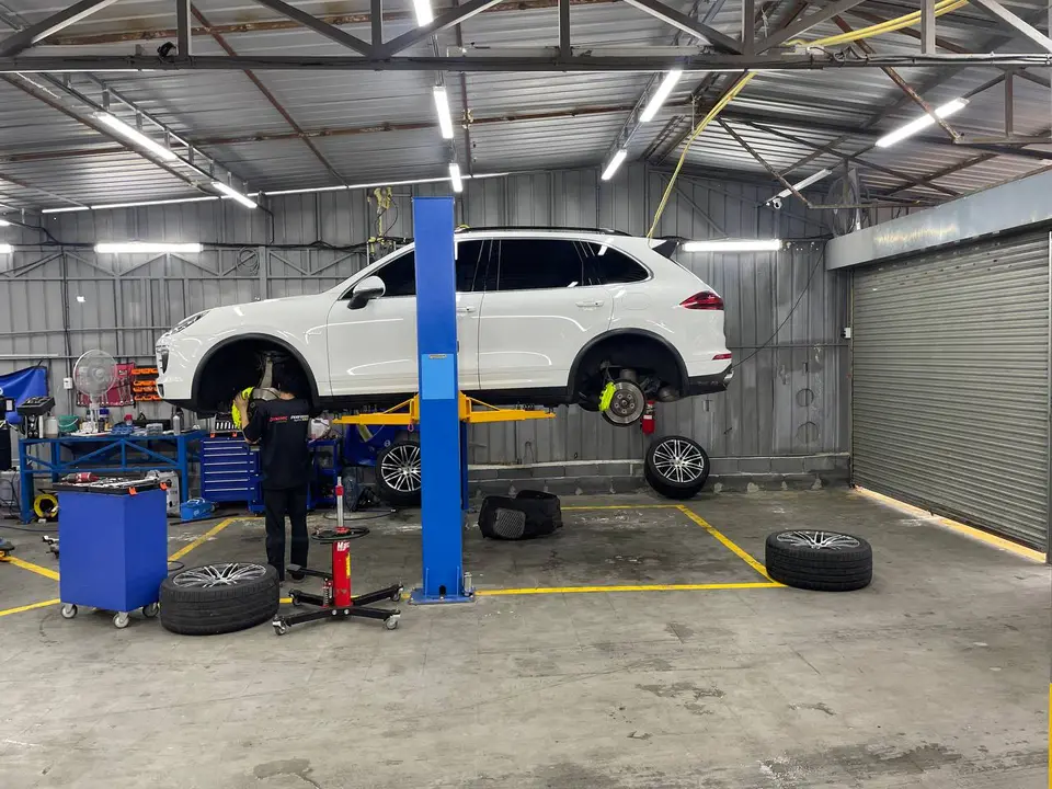 Porsche Cayenne 2020: Белый Porsche Cayenne поднят на подъёмнике, видны снятые колёса и снятый передний правый колесный диск. Работник в черной форме стоит у автомобиля, рядом на полу лежат шины и кол