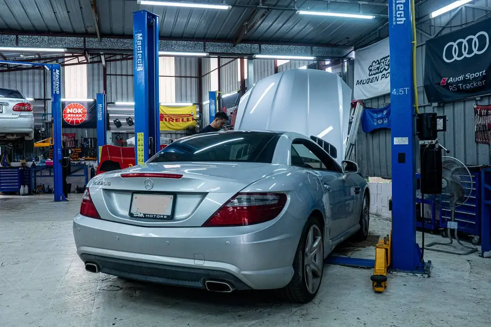 Mercedes-Benz SLC 2017: На фото серебристый Mercedes-Benz SLC 2017, поднятый на подъёмнике в автосервисе. Открыт капот, виден механик, работающий под ним. Видны элементы салона и задняя часть автомоби