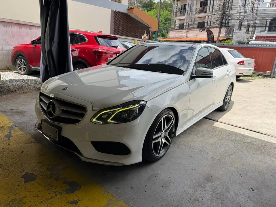 Mercedes-Benz E350 2015: На фото белый Mercedes-Benz E350 2015 года, стоящий на открытой стоянке. Видна передняя часть автомобиля, включая фары, решётку радиатора с логотипом Mercedes-Benz и колёсные 
