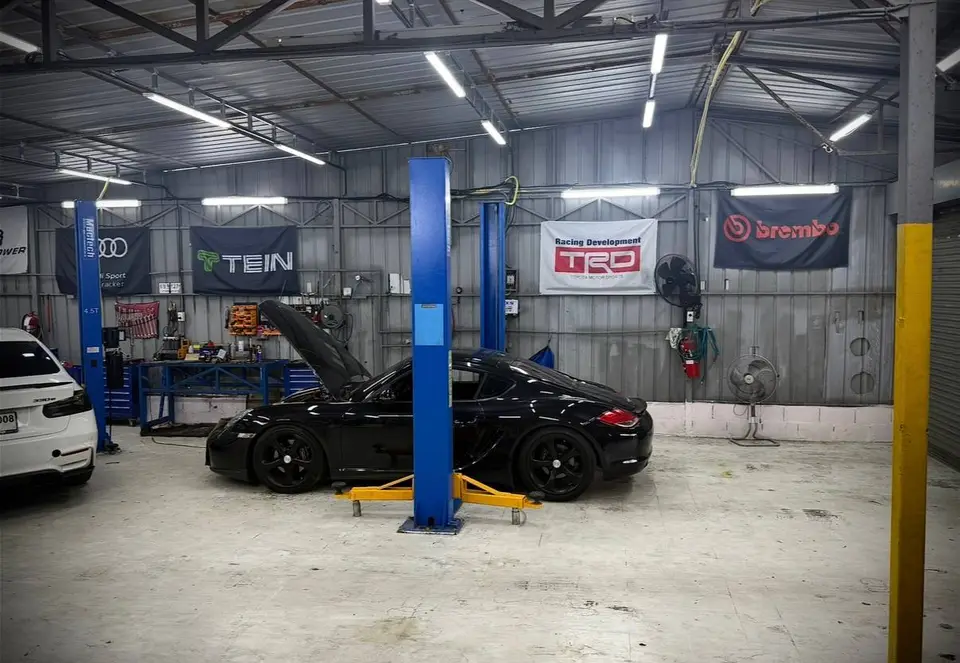 Porsche Cayman 2015: На фото чёрный Porsche Cayman 2015, поднятый на подъёмнике. Капот автомобиля открыт, видна передняя часть кузова и колёсные диски. В гараже также видны инструменты, оборудование и
