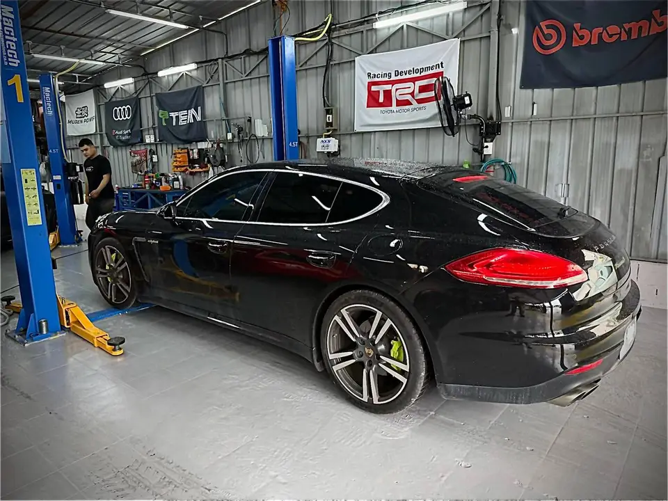 Porsche Panamera 2015: Черный Porsche Panamera 2015 стоит на подъёмнике в автосервисе. На автомобиле установлены тормозные суппорты с зелёной окраской, видны колёсные диски и задние фонари. Вокруг — о