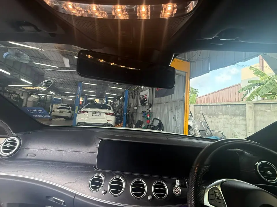 Mercedes-Benz E300d 2020: На фото виден салон автомобиля Mercedes-Benz E300d 2020, снятый изнутри. На переднем плане — приборная панель, руль и центральная консоль. Через лобовое стекло видна автосерв