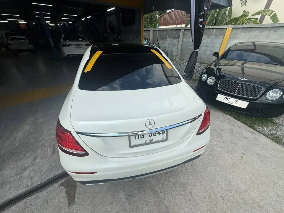 Mercedes-Benz E300d 2020: На фото виден белый седан Mercedes-Benz E300d 2020 сзади. На заднем стекле наклеены желтые ленты, автомобиль находится на подъезде к автосервису. Справа видна часть черного а