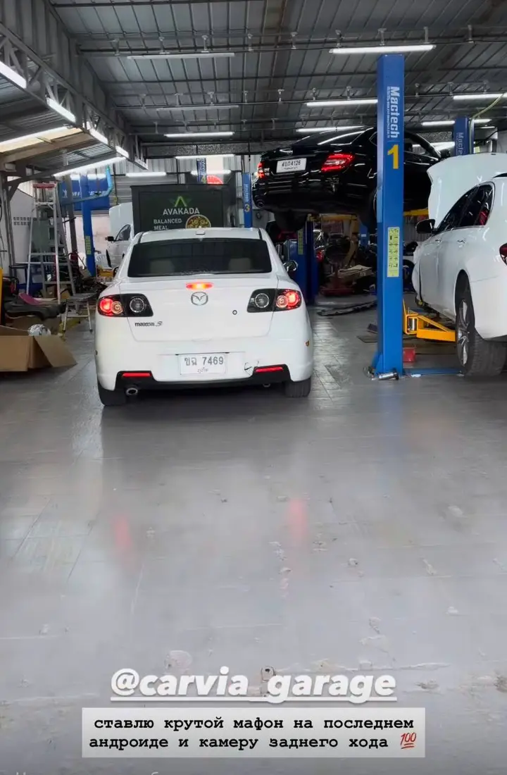 Mazda 3 2013: На фото белый Mazda 3 2013 года, стоящий в гараже. Виден задний бампер, фонари, номерной знак и выхлопная труба. Автомобиль находится на земле, рядом — подъёмник с другим автомобилем.