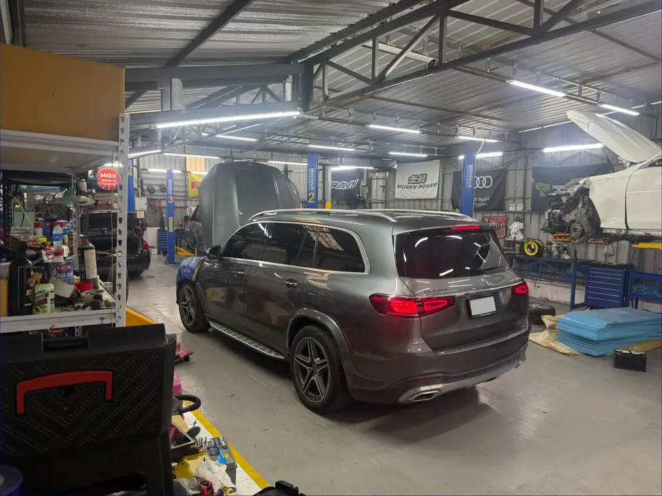 Mercedes-Benz GLS 2023: На фото серебристый Mercedes-Benz GLS 2023, стоящий в гараже. Видна задняя часть автомобиля, открытый багажник и подъёмник с другим автомобилем на заднем плане.