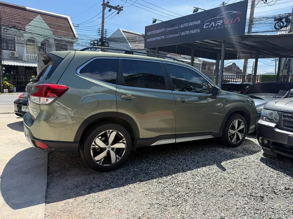Subaru Forester 2022: На фото серый Subaru Forester 2022, стоящий на открытой стоянке автосервиса. Видна боковая часть кузова, колесо с оригинальным дизайном диска, окрашенные в серебристый цвет, и бо
